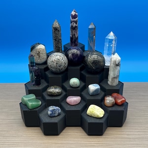 May include: A collection of various polished and raw mineral stones, including spheres, towers, and tumbled pieces, arranged on a tiered black hexagonal display stand. The stones feature diverse colors, textures, and patterns, including clear, purple, grey, green, and red hues.