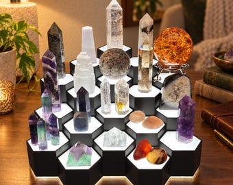 Peut inclure: Un présentoir hexagonal noir à plusieurs niveaux avec éclairage LED intégré, mettant en valeur une collection de divers cristaux, dont des pointes de quartz clair, des tours d'améthyste, une grande sphère en verre craquelé orange et des pierres polies.