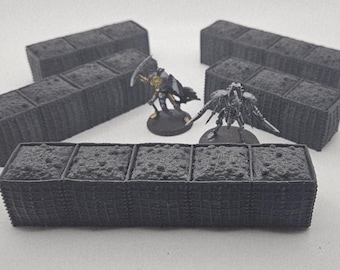 May include: A collection of dark gray, rectangular terrain pieces with textured surfaces, likely for tabletop gaming. Two miniature figures stand nearby. The pieces are divided into sections, suggesting they represent modular structures or containers.