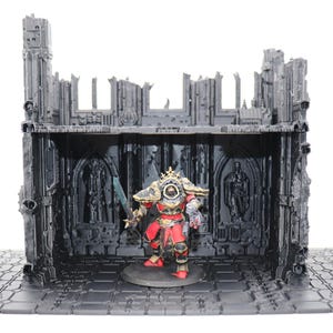 May include: A miniature figure in red and gold armor stands within a detailed, gray, ruined building diorama. The diorama features architectural details and a textured base, suitable for tabletop gaming.