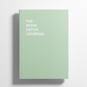 May include: A mint green journal with the text "THE MIND DETOX JOURNAL" in white. The journal is rectangular and appears to be closed, with a shadow cast to the left. The cover is a solid, light green color.