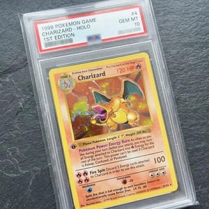 May include: A graded 1999 Pokémon Game Charizard Holo 1st Edition card. The card features a Charizard illustration with a yellow and orange background. The card is encased in a protective holder with a GEM MT 10 grade.
