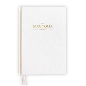 May include: A white journal with the words "DAS MAGNOLIA - TAGEBUCH" in gold lettering. The journal has a textured cover and a ribbon bookmark. The spine is a light beige color.