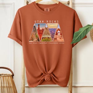 以下が含まれることがあります： さび色のTシャツに、ユタ州の国立公園をそれぞれ描いた5つの三角形が描かれたグラフィックデザインが施されています。三角形の上に「Utah Rocks」という文字が印刷され、その下にザイオン、ブライス、キャピトルリーフ、キャニオンランズ、アーチーズの各公園の名前が記載されています。