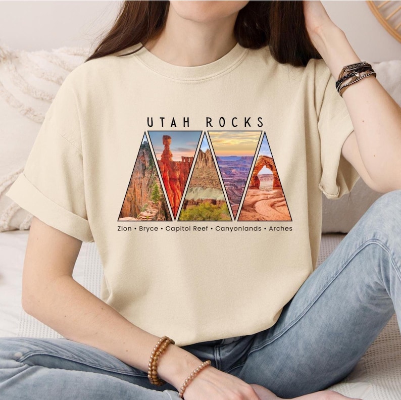以下が含まれることがあります： 前面に「ユタロック」という文字がプリントされたベージュのTシャツ。テキストは、ザイオン、ブライス、キャピトルリーフ、キャニオンランズ、アーチーズなど、ユタ州の国立公園の三角形の画像5つで囲まれています。
