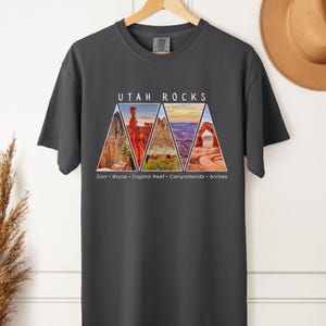 以下が含まれることがあります： 暗いグレーのTシャツに、ユタ州の国立公園の三角形の画像が5つ描かれています。画像の上には「Utah Rocks」という文字が、下には「Zion - Bryce - Capitol Reef - Canyonlands - Arches」という文字が書かれています。