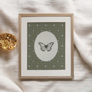 May include: A framed print with a green background and a white oval containing a black and white butterfly illustration. The background has a repeating pattern of small diamonds.