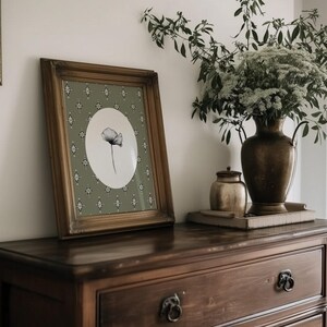 May include: A framed black and white botanical print of a single poppy flower on a green background with a white border. The frame is a dark wood with a distressed finish. The print is sitting on a dark wood dresser with a brass drawer pull.