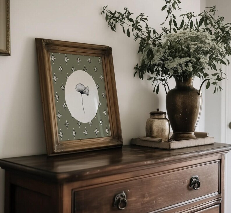 May include: A framed black and white botanical print of a single poppy flower on a green background with a white border. The frame is a dark wood with a distressed finish. The print is sitting on a dark wood dresser with a brass drawer pull.