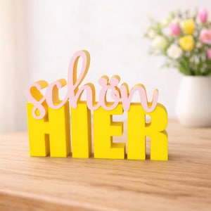 Könnte beinhalten: Ein gelbes Holzschild mit den Worten "schön HIER" in Rosa und Gelb. Das Schild steht auf einem Holztisch, im Hintergrund eine Vase mit Blumen.