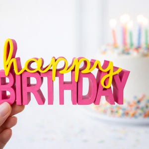 Könnte beinhalten: Ein dreidimensionaler Tortenstecker mit dem Wort happy in gelber Schreibschrift, das über dem Wort BIRTHDAY in kräftigen pinkfarbenen Blockbuchstaben liegt. Der Tortenstecker wird vor einer weiß glasierten Torte mit bunten Streuseln gehalten.