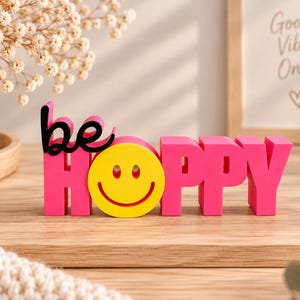 Könnte beinhalten: Ein dreidimensionales Schild mit dem Wort "be" in schwarzer Schreibschrift über dem Wort "HOPPY" in kräftigen, leuchtend pinken Blockbuchstaben, wobei ein gelbes Smiley-Gesicht den Buchstaben "O" im Wort "HOPPY" ersetzt.