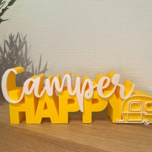 Könnte beinhalten: Gelb-weißes Dekoschild mit der Aufschrift "Camper Happy" und einer Wohnwagen-Grafik. Das Schild besteht aus geschichteten Materialien und ist so konzipiert, dass es auf einer ebenen Fläche aufrecht steht. Das Schild ist eine fröhliche Dekoration.