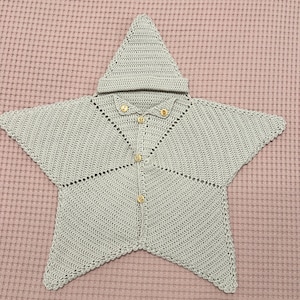 Puede incluir: Un cárdigan para bebé en forma de estrella, de color verde claro, con capucha. El cárdigan de ganchillo hecho a mano tiene cierre de botones y botones dorados. El cárdigan se muestra sobre una superficie rosa texturizada.