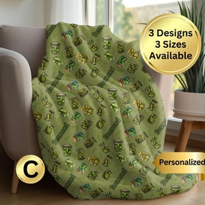 May include: A green fleece throw blanket with a repeating pattern of pickle jars and the name "Desmond" printed on each jar. The blanket is folded and displayed on a chair.