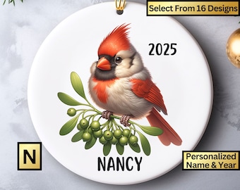May include: A white ceramic Christmas ornament featuring a red cardinal perched on a mistletoe sprig with green berries. The ornament is personalized with the name "NANCY" and the year "2025".