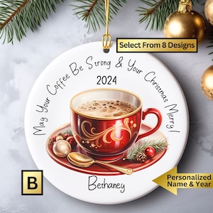 May include: A white ceramic Christmas ornament with a red and gold coffee cup design. The ornament features the text "May Your Coffee Be Strong & Your Christmas Merry!" and the year "2024". The ornament is personalized with the name "Bethany".