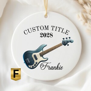 May include: A round white ceramic ornament featuring a blue electric bass guitar graphic. The text CUSTOM TITLE is printed in black above the year 2028, and the name Frankie is printed in a black script font below the guitar. A gold string is attached.