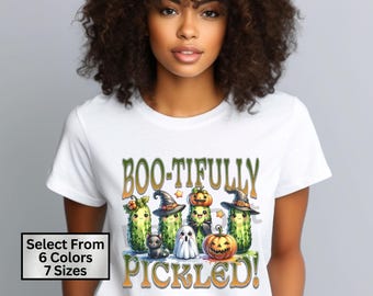 May include: White t-shirt with a green, orange, and black graphic of cartoon pickles dressed as witches and ghosts. The text "Boo-tifully Pickled!" is printed on the shirt.