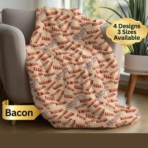May include: A beige blanket with a repeating pattern of bacon strips and the name "Abigale" in a script font. The blanket is draped over a chair. Text on the image reads "4 Designs 3 Sizes Available" and "Bacon".