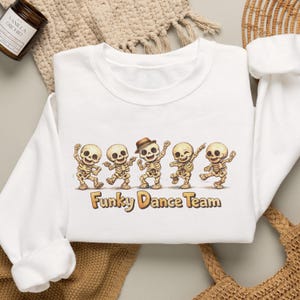 May include: A white long-sleeve sweatshirt featuring a graphic of five dancing skeletons. The center skeleton wears a brown fedora hat. Below the skeletons, the text reads Funky Dance Team in a bold, stylized brown font with a light outline.