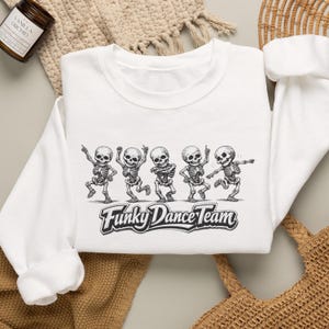 May include: A white crewneck sweatshirt featuring a black graphic of five skeletons in various dancing poses. Below the skeletons, the text reads Funky Dance Team in a bold, stylized font. The sweatshirt has long sleeves and a ribbed neckline.