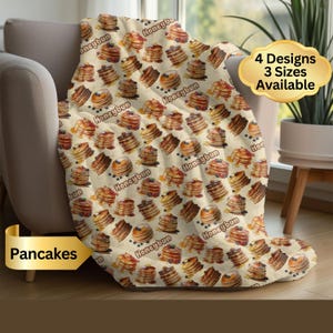 May include: A beige blanket with a repeating pattern of pancakes, blueberries, and syrup. The blanket has the text "Honeybun" and "Pancakes" on it. The blanket is available in 4 designs and 3 sizes.