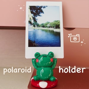 May include: A green frog figurine holding a white Polaroid photo frame. The frog is sitting on a red and white mushroom-shaped base. The photo frame has a picture of a lake with a fishing line in the water.