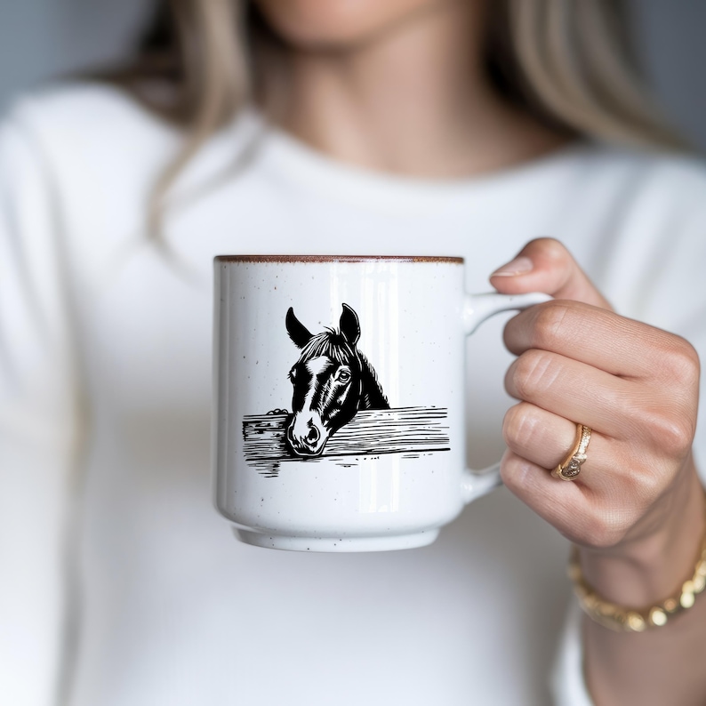 May include: White ceramic mug with a brown rim featuring a black and white illustration of a horse's head peeking over a wooden fence. The mug has a large handle and a speckled texture.
