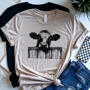May include: Beige t-shirt featuring a black and white illustration of a cow peering over a wooden fence. The shirt is folded, with a black t-shirt and blue jeans visible in the background. A phone with a black and white checkered case is also visible.