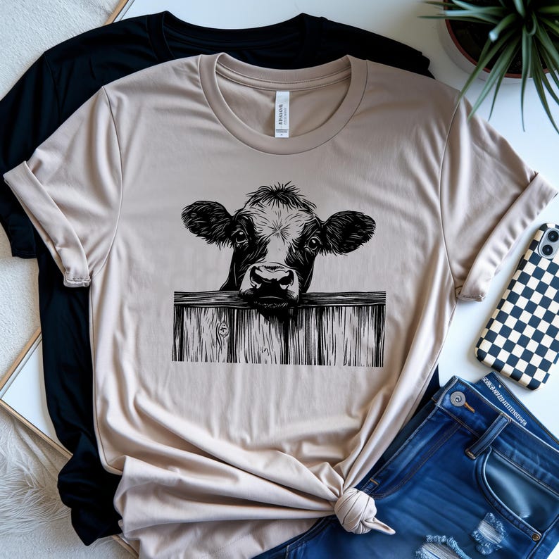 May include: Beige t-shirt featuring a black and white illustration of a cow peering over a wooden fence. The shirt is folded, with a black t-shirt and blue jeans visible in the background. A phone with a black and white checkered case is also visible.