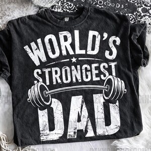 May include: Black t-shirt with the text "WORLD'S STRONGEST DAD" in white, featuring a barbell graphic. The shirt has a distressed, vintage look. A star is above the word "STRONGEST".