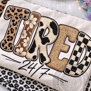 May include: Cream-colored shirt with the word "TIRED" in a mix of animal print, floral, and checkered patterns. The text "24/7" is written below in black script. Leopard print fabric is visible in the background.
