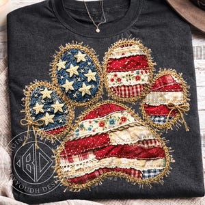 Könnte beinhalten: Dunkelgraues T-Shirt mit einem Pfotenabdruck-Design aus Stoffflicken. Die Flicken sind rot, weiß und blau, mit Sternen auf den blauen Flicken. Der Pfotenabdruck ist mit goldfarbenem Faden umrandet.