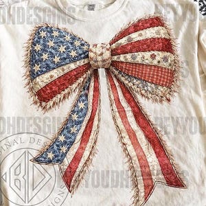 May include: A cream-coloured t-shirt with a large bow design in the style of the American flag. The bow features red and white stripes, blue stars, and a vintage, textured appearance. The shirt has short sleeves and a relaxed fit.