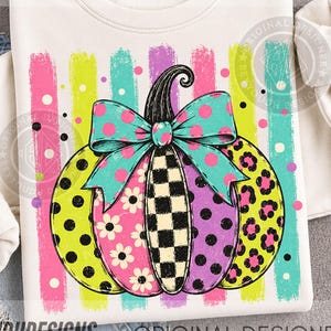 May include: A cream-coloured sweatshirt featuring a graphic of a pumpkin with multicoloured segments in polka dot, floral, checkered, and leopard patterns. A large teal bow with pink polka dots sits atop the pumpkin, set against vertical stripes.