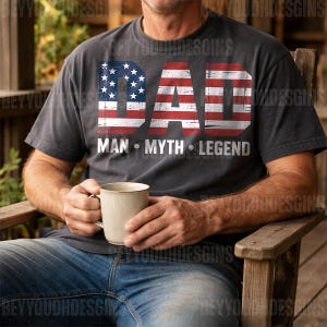 Puede incluir: Camiseta gris oscuro con la palabra "DAD" en un dise&ntilde;o de bandera americana desgastada. Debajo, las palabras "MAN &bull; MYTH &bull; LEGEND" en blanco. La persona sostiene una taza beige.