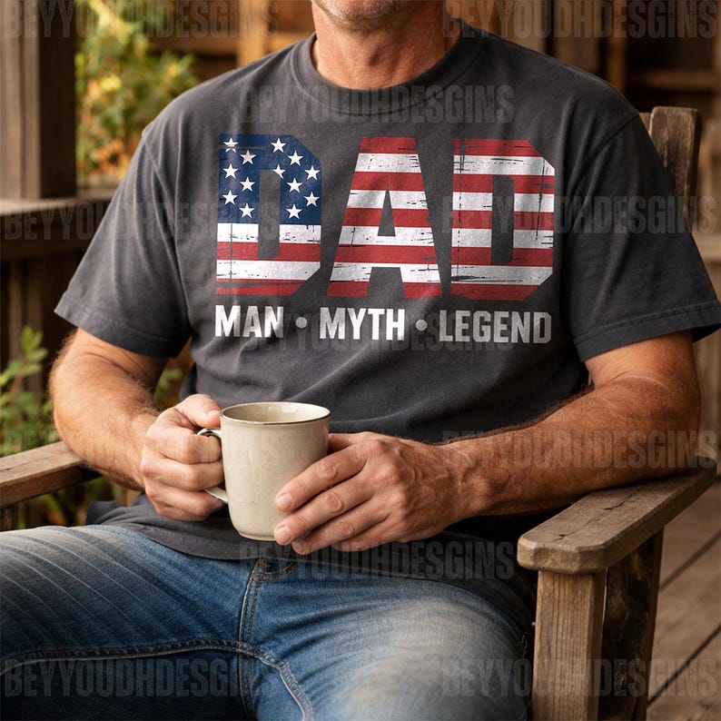 Puede incluir: Camiseta gris oscuro con la palabra "DAD" en un dise&ntilde;o de bandera americana desgastada. Debajo, las palabras "MAN &bull; MYTH &bull; LEGEND" en blanco. La persona sostiene una taza beige.