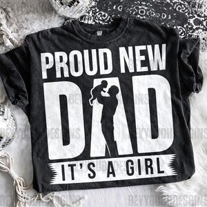 Puede incluir: Camiseta negra con el texto "PROUD NEW DAD" en letras blancas grandes. Debajo de la palabra "DAD" hay una silueta de una persona sosteniendo a un beb&eacute;. El texto "IT'S A GIRL" est&aacute; impreso en blanco en la parte inferior.