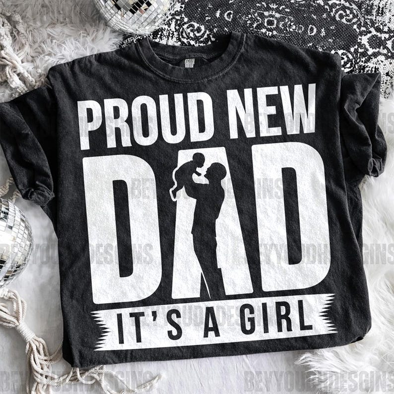 Puede incluir: Camiseta negra con el texto "PROUD NEW DAD" en letras blancas grandes. Debajo de la palabra "DAD" hay una silueta de una persona sosteniendo a un beb&eacute;. El texto "IT'S A GIRL" est&aacute; impreso en blanco en la parte inferior.