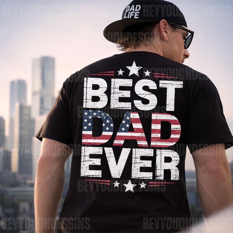 Może przedstawiać: Czarny t-shirt z białym napisem "BEST DAD EVER", z napisem "DAD" wypełnionym wzorem amerykańskiej flagi. Koszulka ma r&oacute;wnież gwiazdy i paski. Widoczna jest czarna czapka z napisem "DAD LIFE".