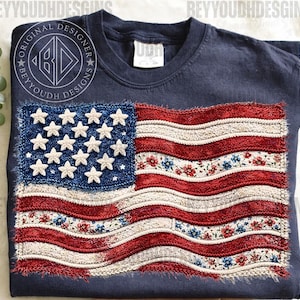 May include: Navy blue t-shirt featuring a distressed American flag design. The flag has a blue field with white stars and red and white striped sections, some with floral patterns. The shirt has the text "BEYYOUHDESIGNS" around a circular logo.
