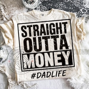 Puede incluir: Camiseta color crema con el texto "STRAIGHT OUTTA MONEY" en negro desgastado, enmarcado por un rect&aacute;ngulo negro. Debajo, el hashtag "#DADLIFE". Es una prenda de vestir informal.