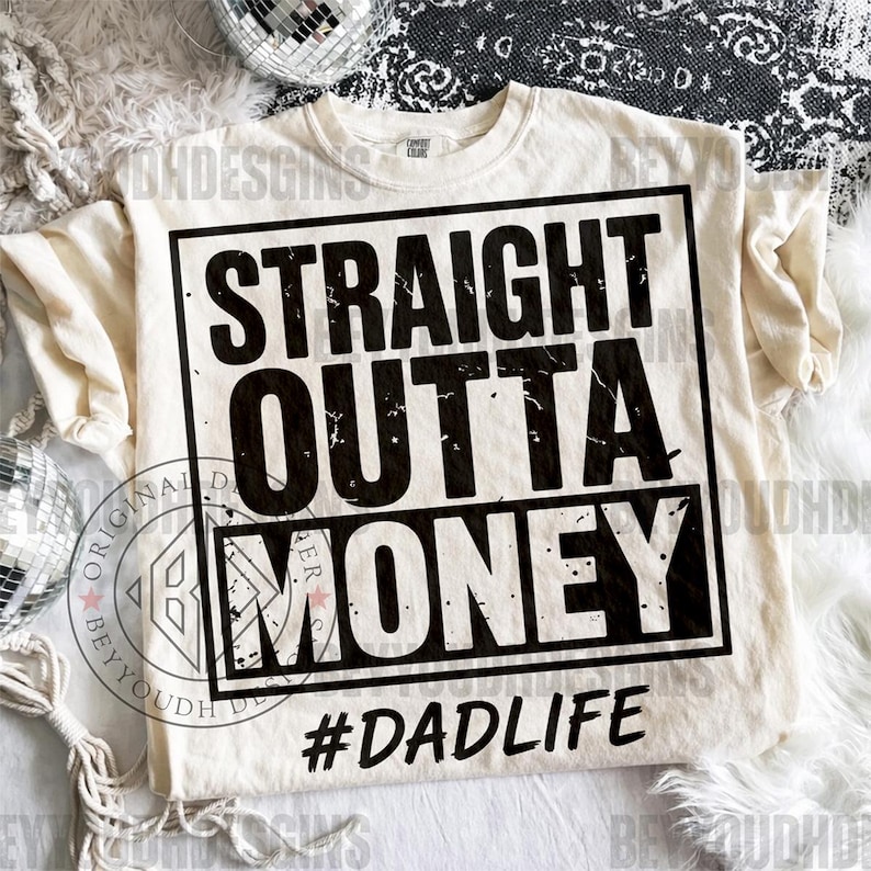 Puede incluir: Camiseta color crema con el texto "STRAIGHT OUTTA MONEY" en negro desgastado, enmarcado por un rect&aacute;ngulo negro. Debajo, el hashtag "#DADLIFE". Es una prenda de vestir informal.