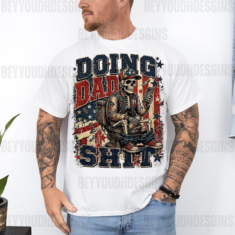 May include: White t-shirt with a graphic design featuring a skeleton wearing a baseball cap and jacket, seated on a toilet, holding a book. The text reads "DOING DAD SHIT" in red, white, and blue, with a distressed American flag background.