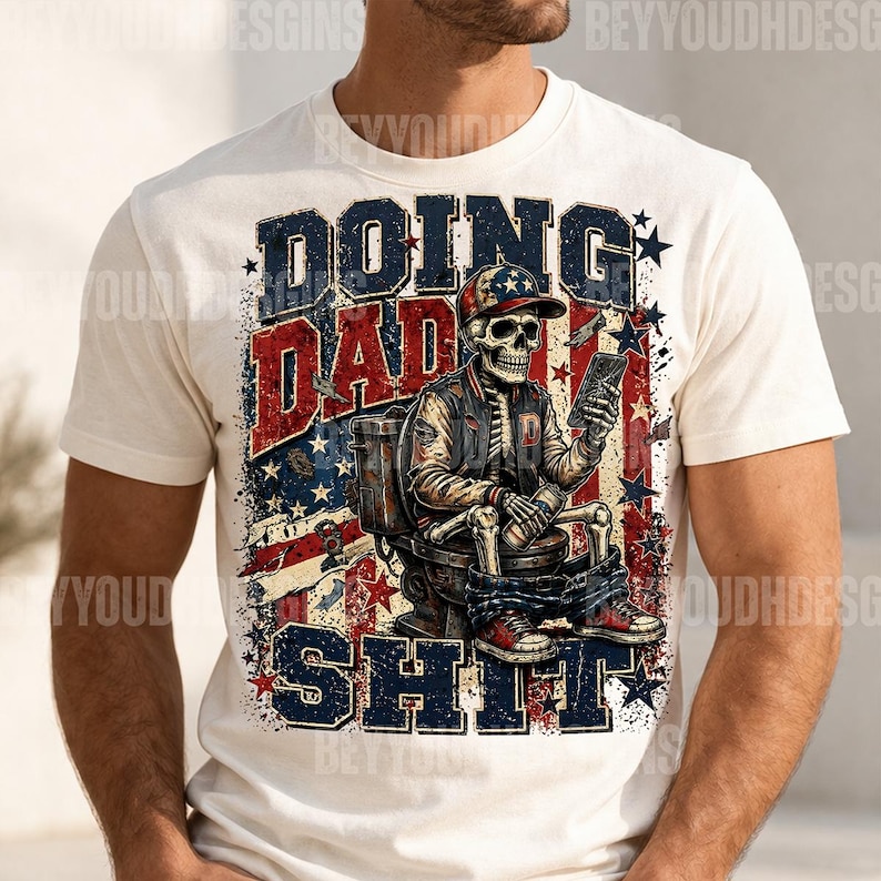 May include: Cream-colored t-shirt with a graphic featuring a skeleton wearing a baseball cap and jacket, seated on a toilet, holding a phone. The text reads "DOING DAD SHIT" in distressed red and blue lettering with star accents.