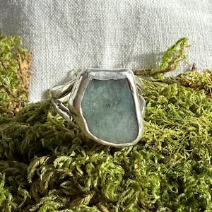 May include: A silver ring with a large, irregular, pale blue-green stone. The ring is set against a backdrop of green moss and a light-colored, textured fabric.