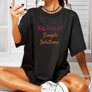May include: Black t-shirt with the text "Big Fan of Simple Solutions" in pink and orange. The shirt is paired with white sneakers and a white purse. The person is holding a smartphone.