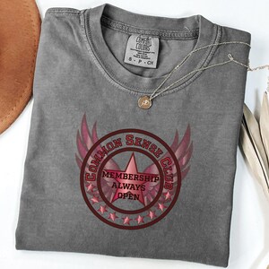 Puede incluir: Una camiseta gris Comfort Colors con un gráfico redondo que dice "COMMON SENSE CLUB MEMBERSHIP ALWAYS OPEN". El gráfico presenta una estrella rosa, alas y estrellas. Un collar dorado descansa sobre la camisa.
