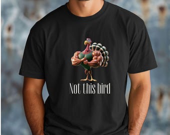 Puede incluir: Camiseta negra con un pavo de dibujos animados con músculos y una cola colorida. El texto "Not this bird" está debajo. La camiseta es de cuello redondo.